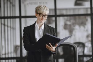 Personne aux cheveux courts blonds portant des lunettes et un tailleur noir, consultant un dossier professionnel dans un espace de bureau moderne.